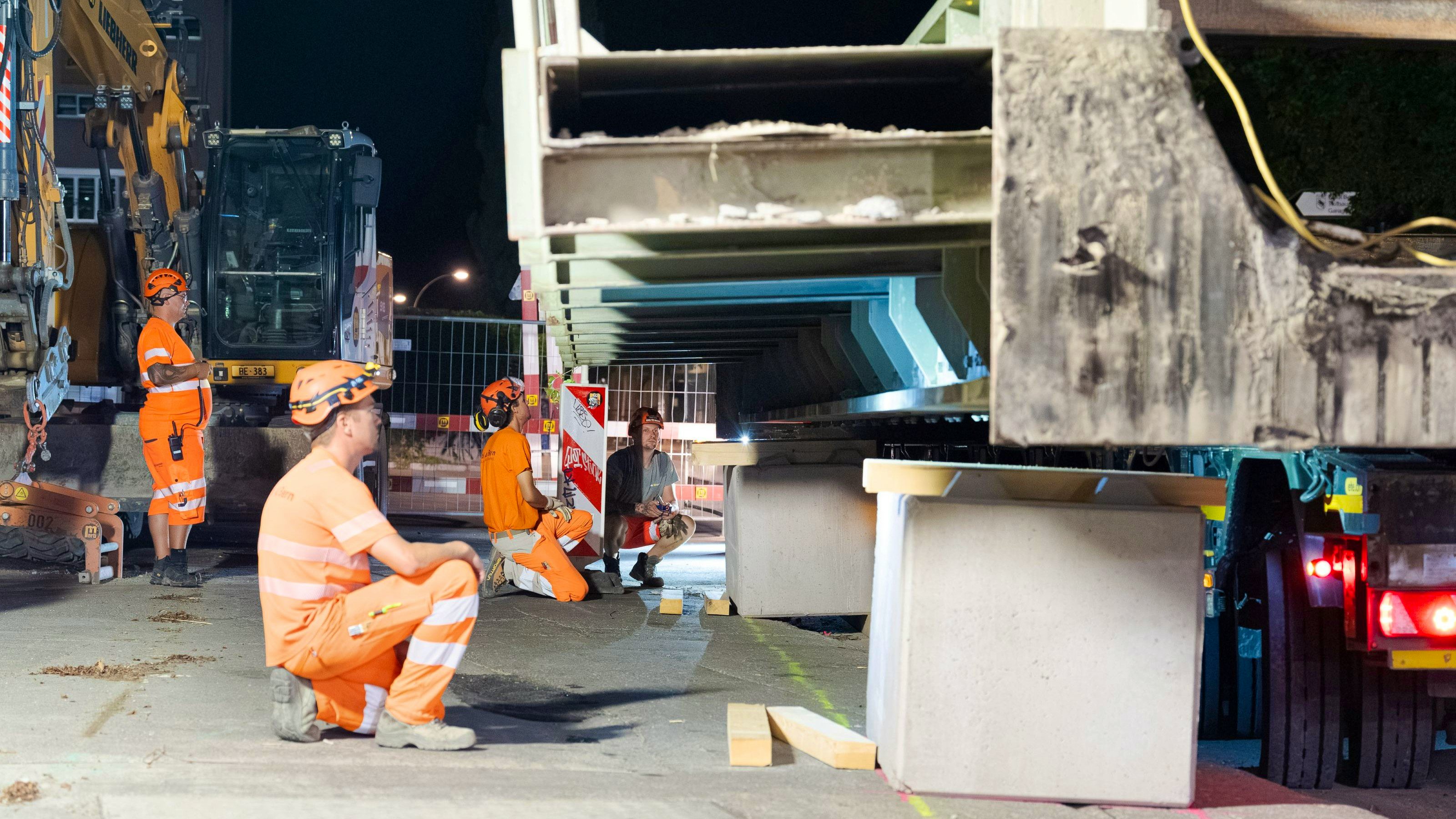 Baustellenszene bei Nacht: Mehrere Bauarbeiter in orangener Schutzkleidung beobachten und positionieren einen ausgehobenen Betonbrückenträger, der auf Abstützungen und einem Tieflader liegt. Im Hintergrund ist ein Bagger und eine Beleuchtung zu sehen.