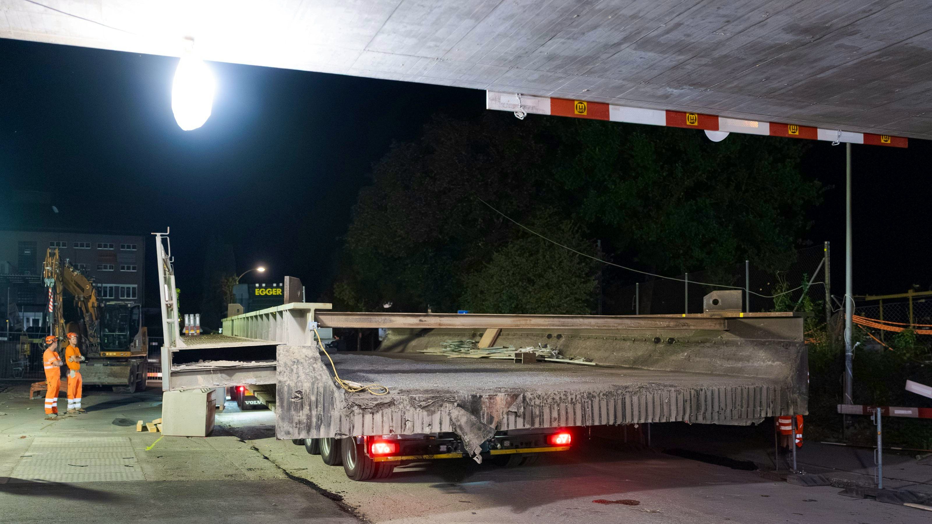 Blick von hinten auf einen grossen Betonbrückenträger, der auf einem Tieflader transportiert wird; am linken Bildrand stehen zwei Arbeiter in Warnkleidung. Die Baustelle ist nachts beleuchtet, im Vordergrund ist die abgesenkte Fahrbahn sichtbar.