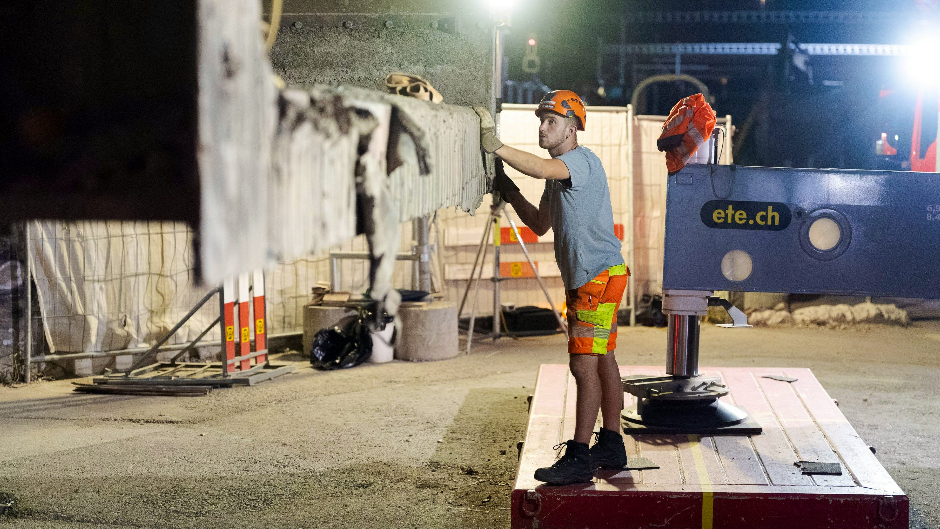 Nahaufnahme eines Arbeiters mit Schutzhelm, der an der Kante eines ausgehobenen Betonträgerteils arbeitet und dieses prüft. Im Hintergrund Baustellenabsperrungen, Beleuchtung und Hebeausrüstung.