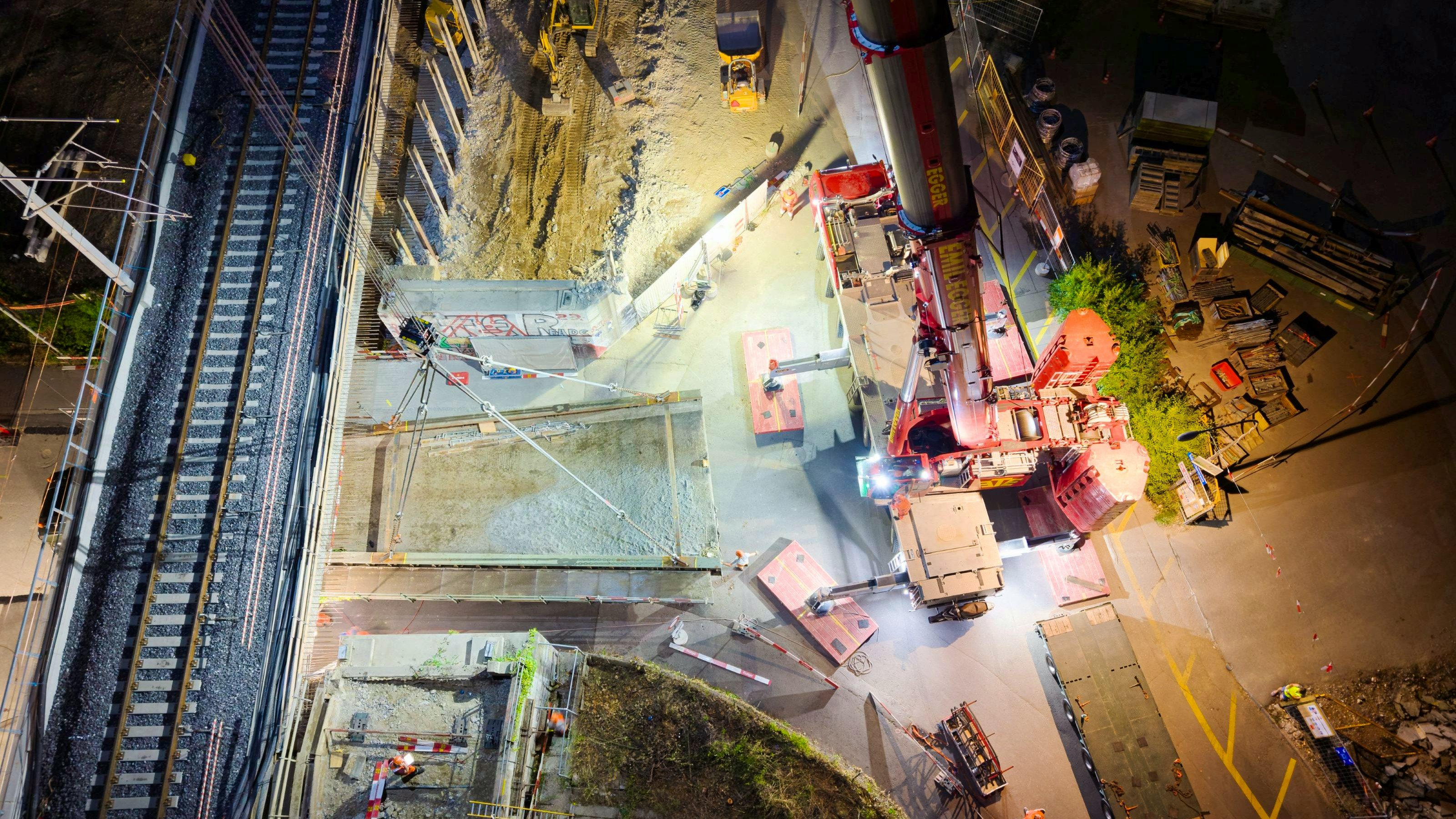 Nachtaufnahme aus der Vogelperspektive: Ein grosser Mobilkran und Hebevorrichtungen stehen auf der Strasse, neben den Gleisanlagen ist eine Baugrube sichtbar. Baustellenleuchten und Markierungen beleuchten mehrere Arbeiter und die Umgebung während des Hebens eines Brückenteils.