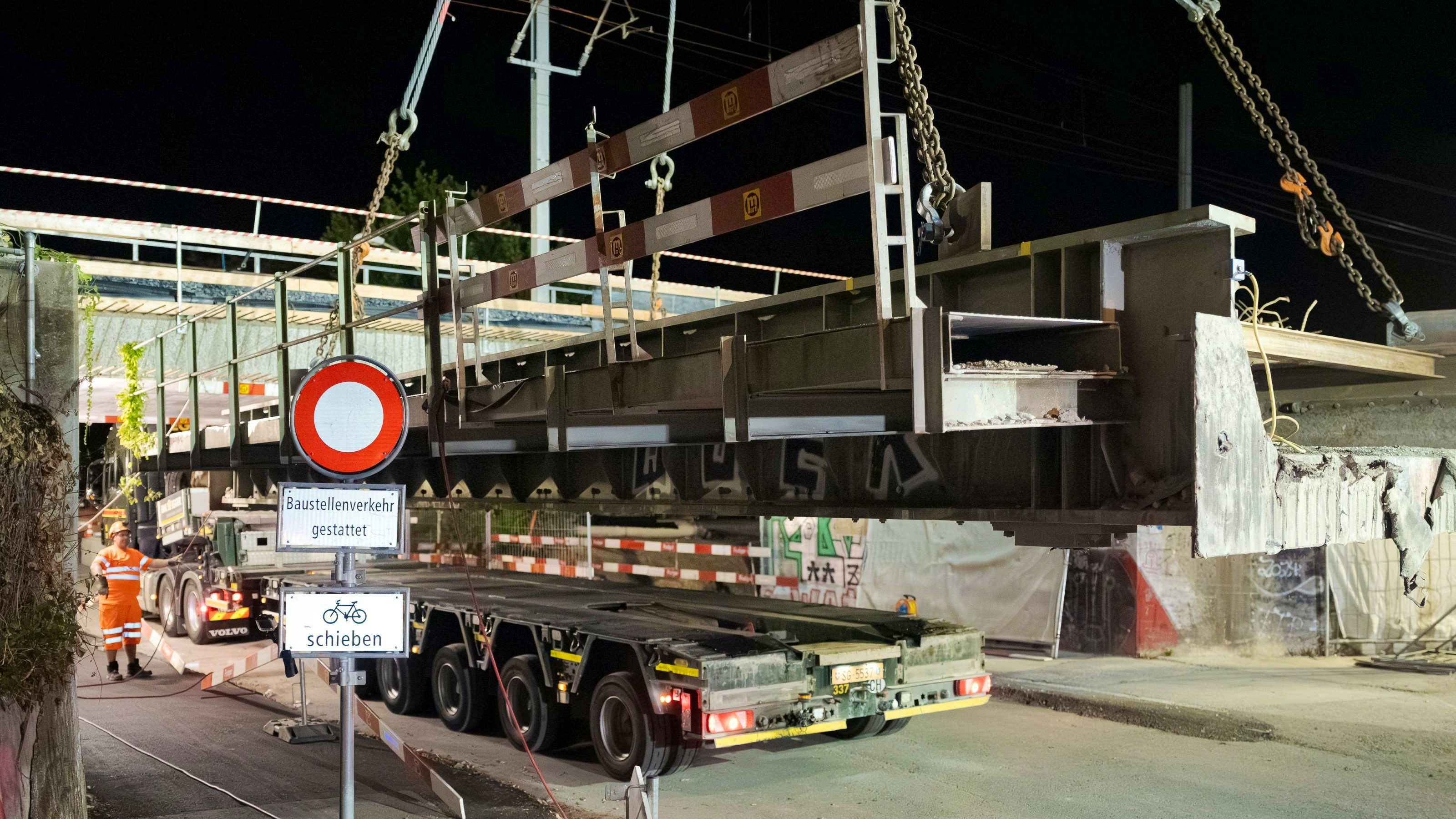 Seitenansicht eines schweren Stahl- oder Betonträgers, der mit Ketten am Kran hängt und gerade über einen Tieflader abgesetzt wird. Ein Verkehrsschild und Absperrungen markieren die Baustelle; im Hintergrund stehen Arbeiter in Warnkleidung.