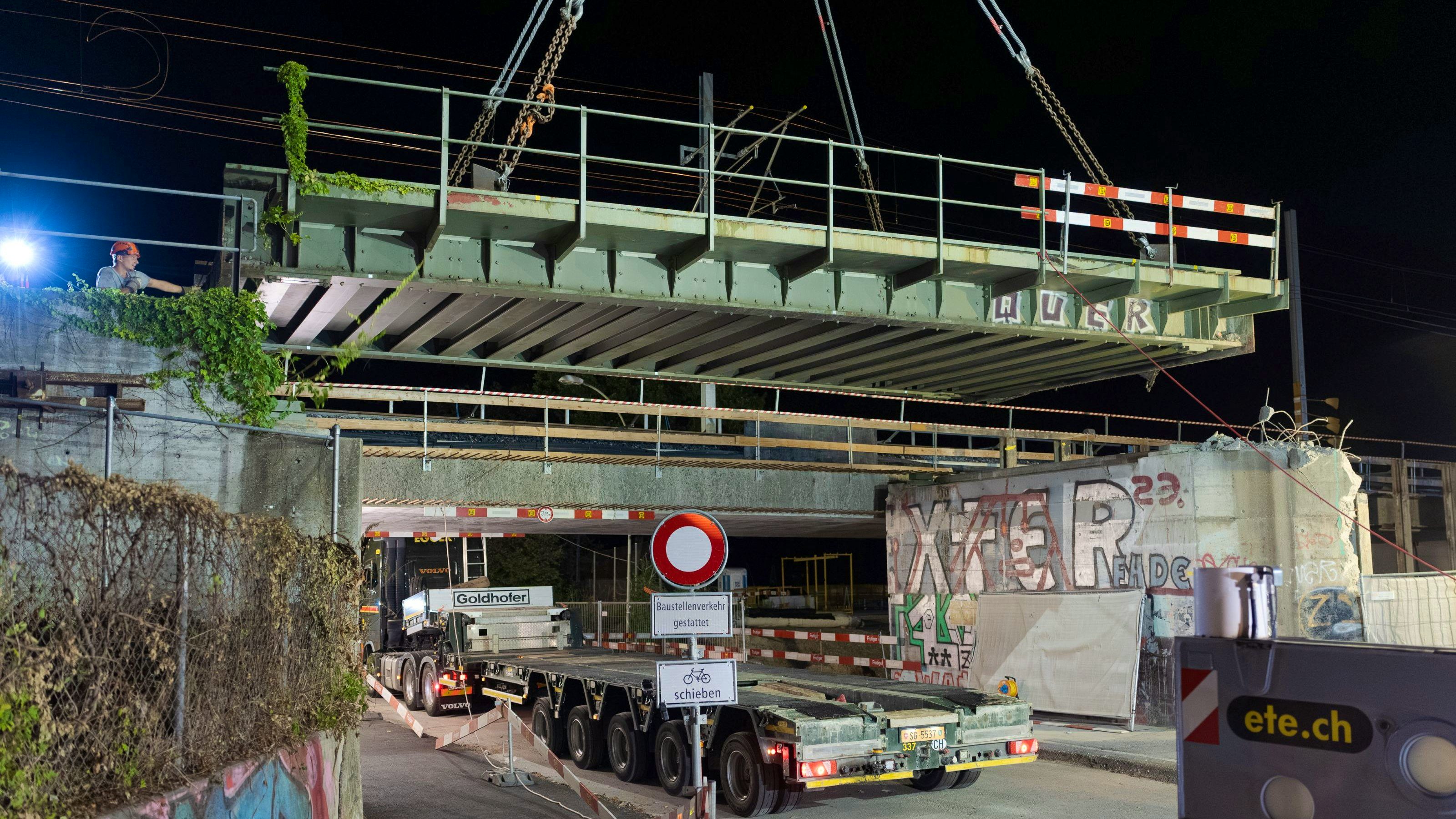 Detailaufnahme des abgesenkten Brückenträgers, der am Kran hängt; zwei Hebeseile sind sichtbar. Im Vordergrund ein Teil des Tiefladers und Baustellenmaterial, im Hintergrund Absperrungen und Graffiti an den Brückenwänden.