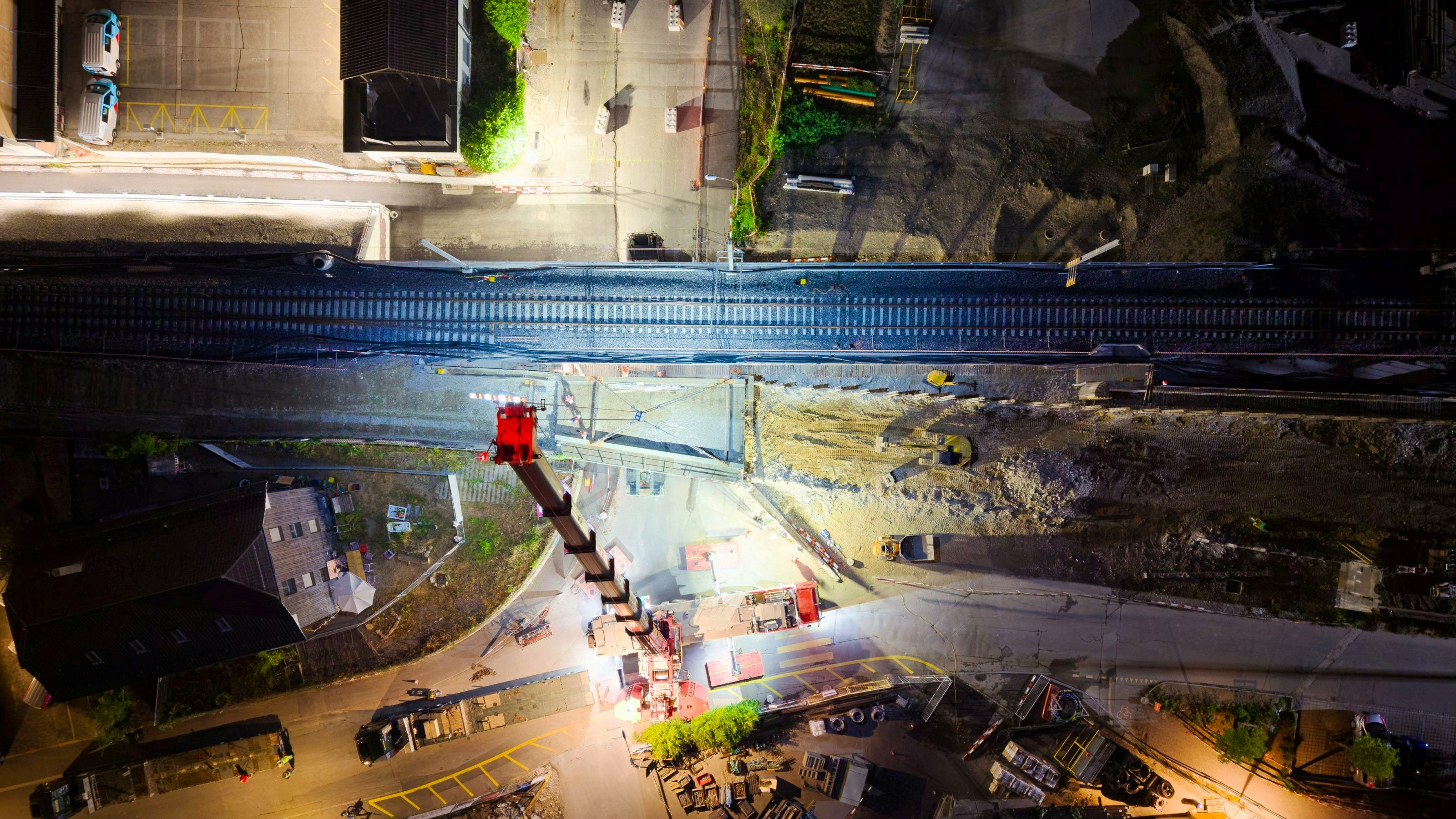Weitwinkel-Nachtaufnahme der Baustelle Stöckacker: Ein grosser Mobilkran hebt ein Brückenteil über der gesperrten Strasse nahe den Bahngleisen. Baustellenbeleuchtung, Baufahrzeuge und vereinzelte Arbeiter sind um den Kran herum sichtbar; im Hintergrund zeigt sich die Stadt bei Nacht.
