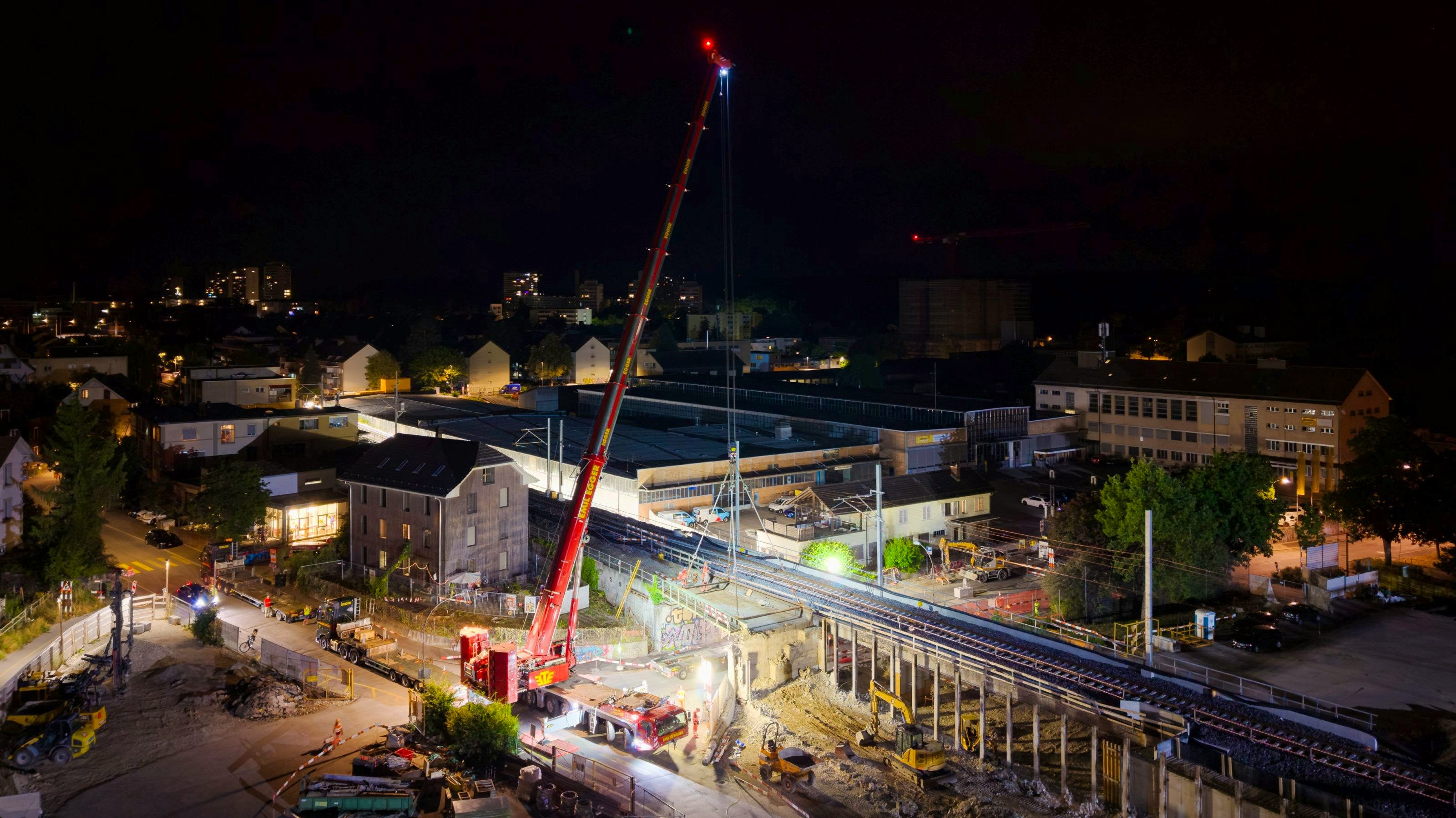 Drohnenaufnahme von oben: Der Mobilkran steht unter dem Bahntrassee und hebt ein Brückenelement, das quer über die Strasse liegt. Gut erkennbar sind die Gleise, Baustellenfahrzeuge, Materiallager und die beleuchtete Arbeitszone in der Nacht.