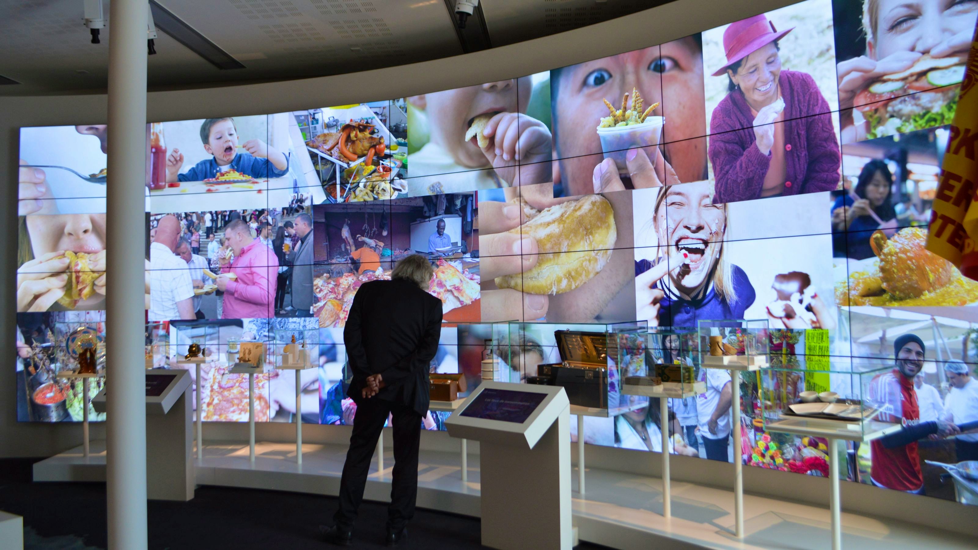 A man stands in front of a large screen which shows various images of people interacting with food.