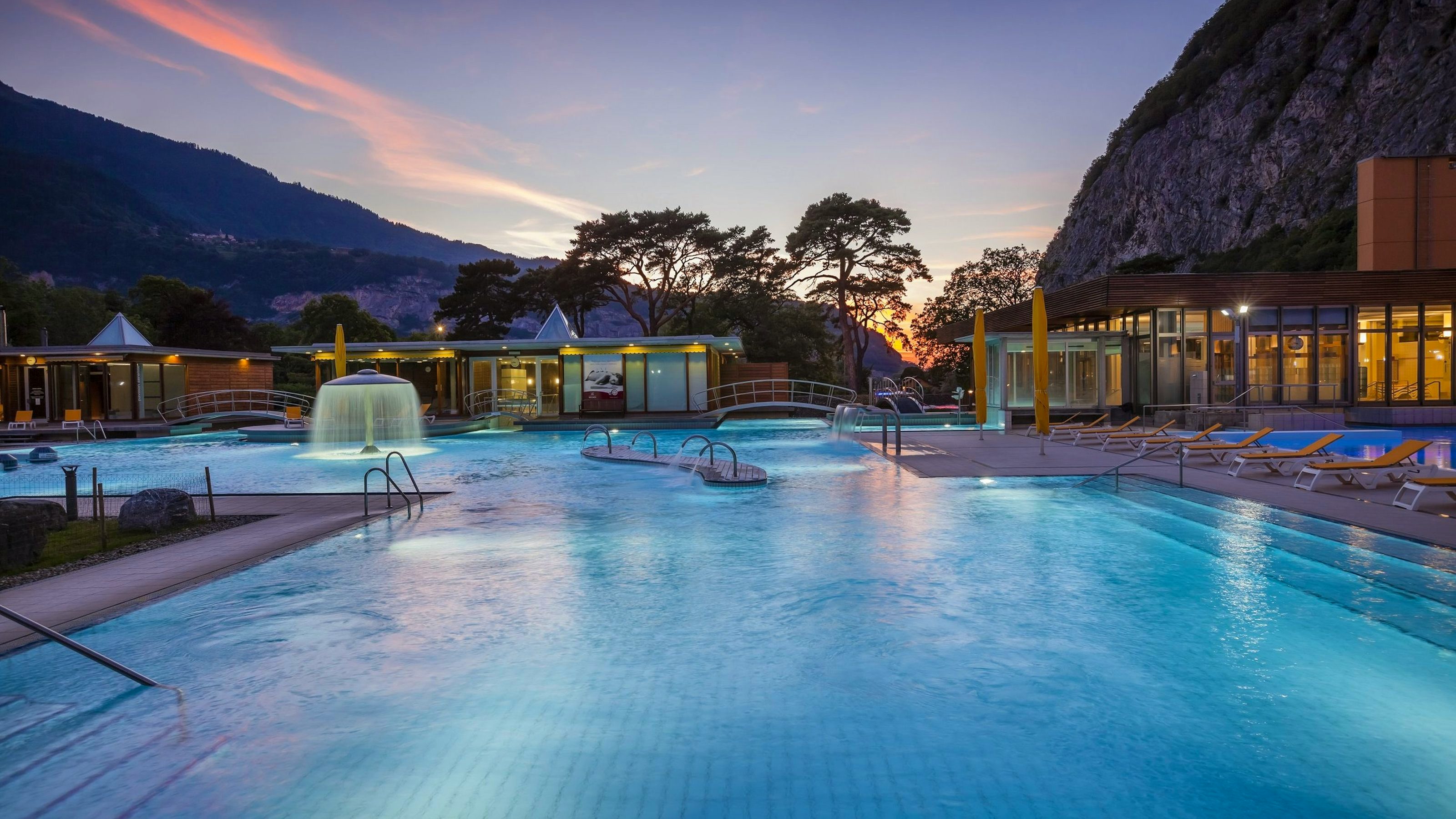 Piscine extérieure d'un centre thermal en soirée, avec une grande fontaine éclairée au centre, plusieurs ponts arqués enjambant les bassins et des chaises longues disposées le long de la piscine. Le site est entouré de montagnes et d’arbres avec un ciel coloré par le coucher du soleil.