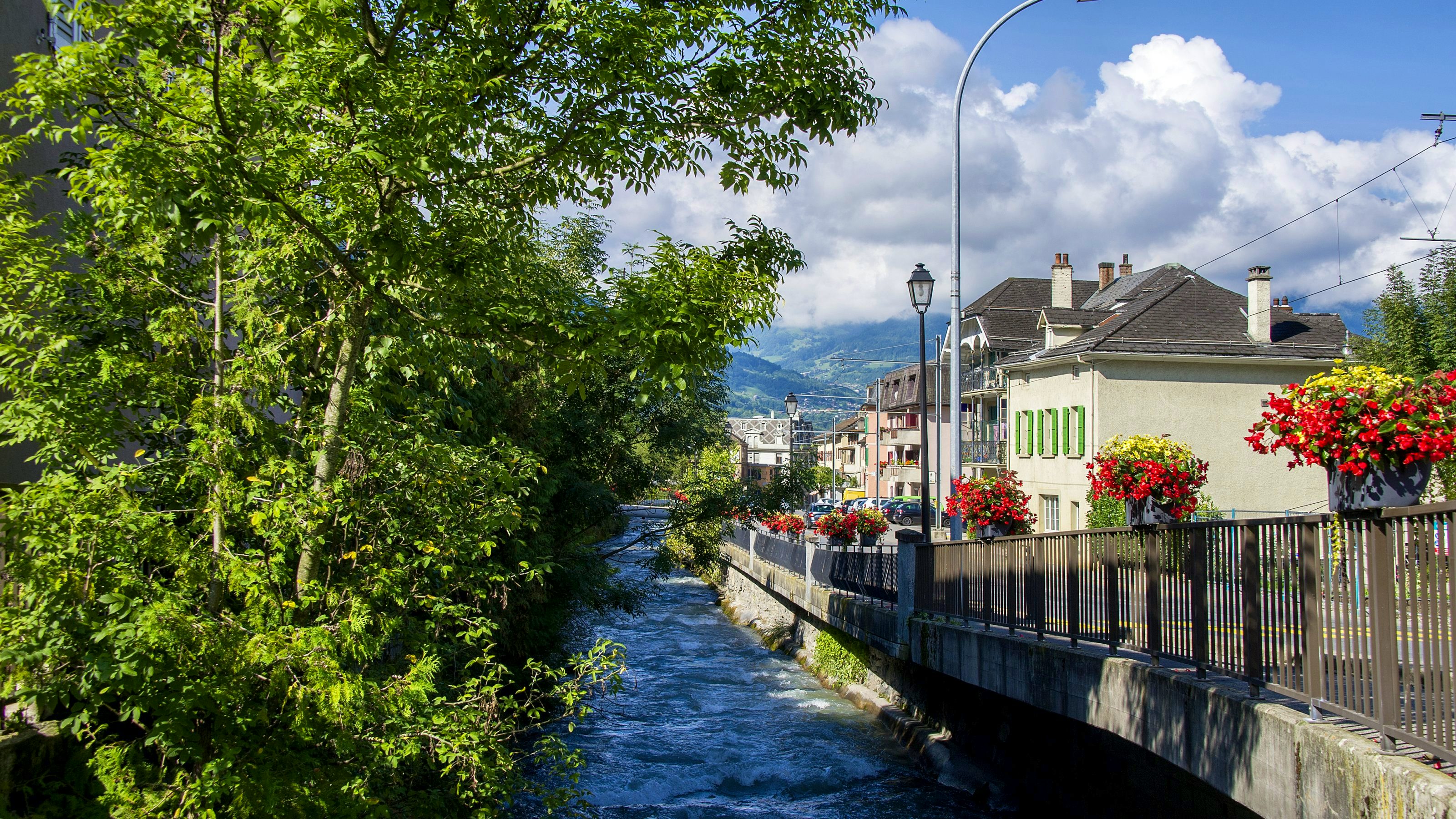À gauche de l’image, des arbres donnent leur ombre à un canal. À droite, une route bordée de maisons aux murs blancs et volets verts. En arrière-plan, on devine des montagnes surplombées de quelques nuages et un ciel bleu.