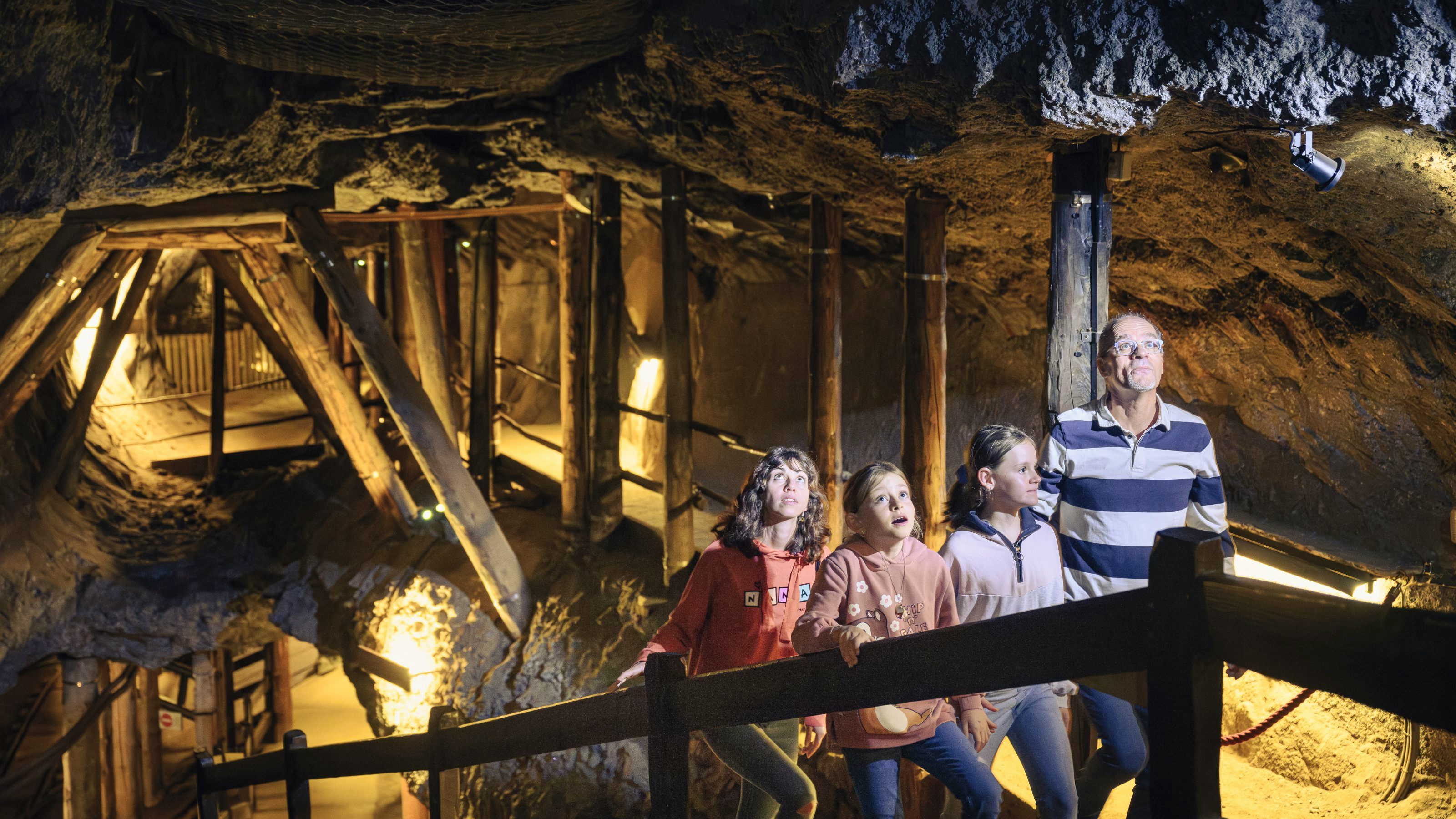 Une famille de quatre personnes explore une galerie souterraine éclairée. Autour d’elles, des parois rocheuses et des poutres en bois qui soutiennent le plafond de la mine. Les enfants et les adultes regardent vers le haut, fascinés par ce qu’ils découvrent.