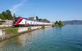 Der FV-Dosto fährt an einem sonnigen Tag am Zürichsee in Richterswil vorbei. Der See befindet sich auf der linken Seite des Zuges.
